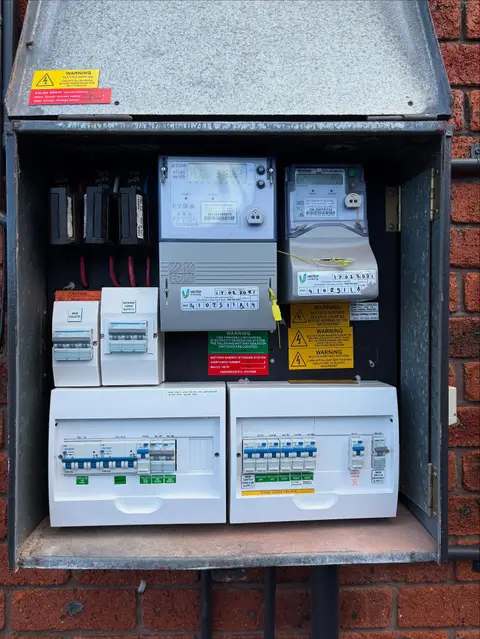 A home electrical switchboard in a wall box with meters and circuit breakers, where a battery system connects