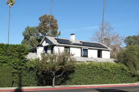 Solar panels on the roof of a white house behind a trimmed hedge